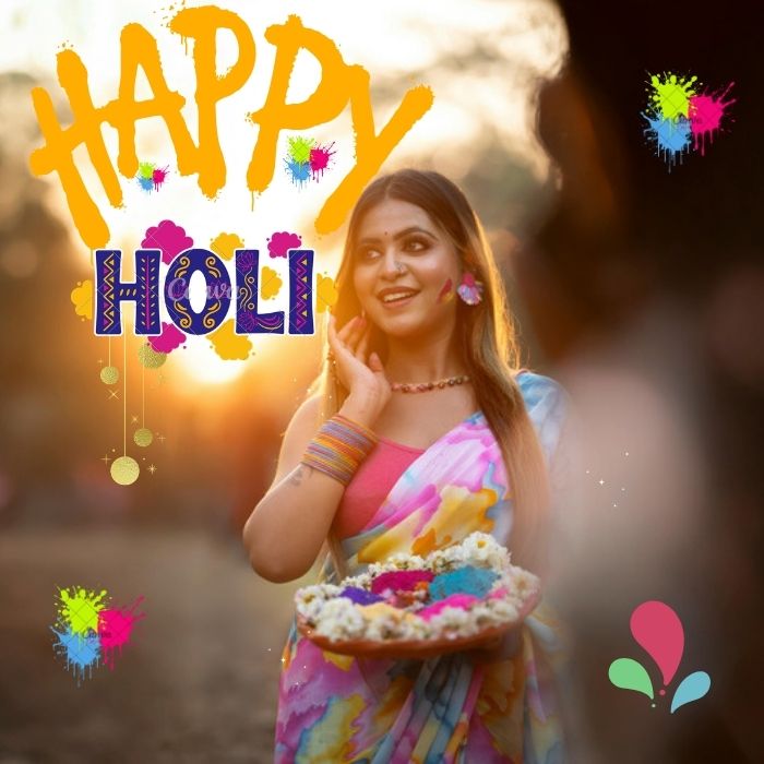 A woman in a colorful saree holds a plate of Holi powders, with “Happy Holi” text and paint splashes overlayed on the image.Happy Holi images.
